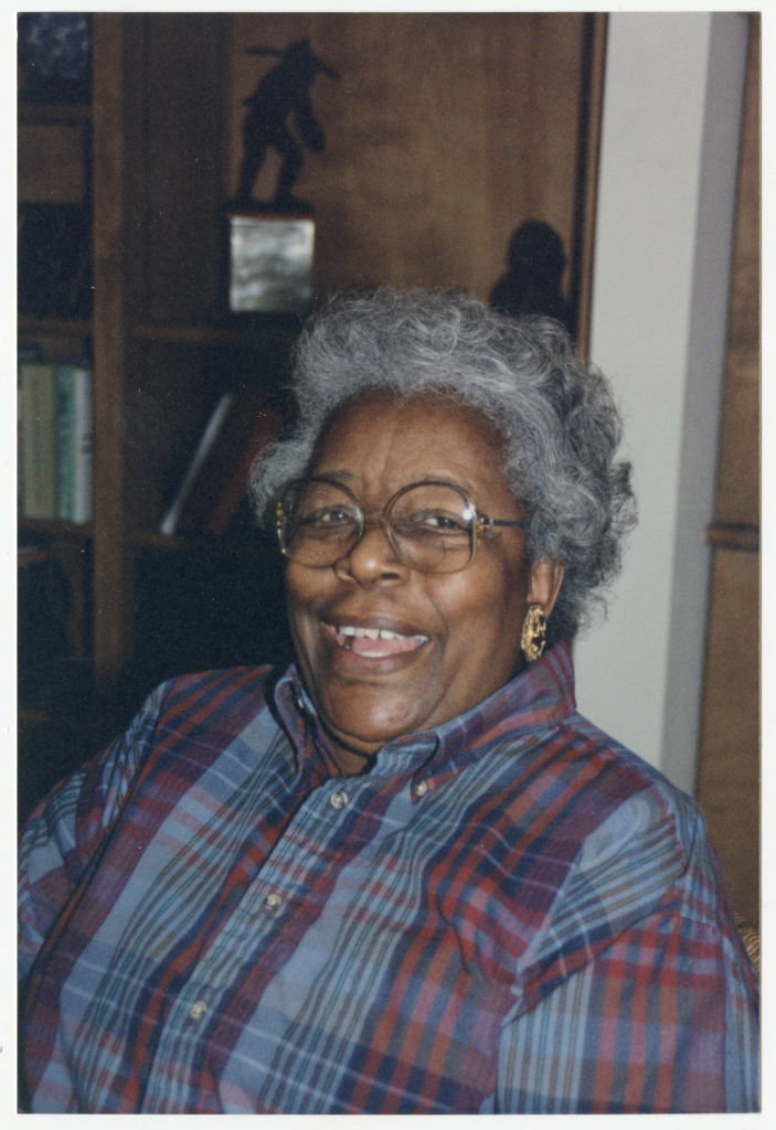 Photo of Bernice Sills Britt. She is seated in front of a bookshelf filled with books, a figurine with plaque, and other memorabilia and knickknacks. She has a wide smile, perhaps she was laughing in this photo. Her hair is greyed and curly. She wears a blue and red plaid button up shirt, large gold earrings with red jewels, and brown, large, circular glasses.