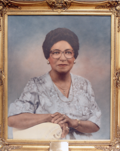 Framed photo of Emma Perry Boyer. She is posed for the photo with a soft blue and pink watercolor backdrop, left arm propped up on a pedestal, right hand folded over her left. She is wearing a gray and white floral short-sleeved shirt. Her hair is curled and she has make-up, pearl earrings, necklace, watch, and rings on. She wears large, circular tortoise shell glasses. There is a small plaque at the bottom of the photo frame with her name and her status as the director of the YWCA.