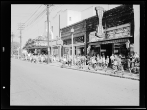 Photo is taken from one side of a street, showing a series of businesses on the other side of the street with Center Theater towards the end on the left side of the photo. The sidewalk is filled with white people, mainly children, heading to the theater. There are bikes parked all along the sidewalk. Towards the back side of the theater, some Black children are seen with placards waiting to the side while white patrons crowd around the theater entrance.