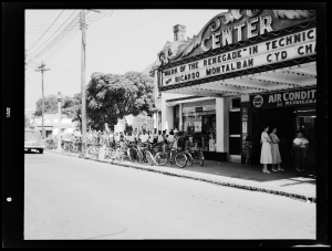 Center theater is pictured with the front in the right half of the frame of the photo. The concession stand is visible, along with an entrance that has a sign hung above it reading “Colored Balcony Entrance.” Outside of the entrance, a group of Black children are waiting and some are posing for the photo, smiling, and holding signs that read “Penney’s Back to School Movie Party.” Some white children are seen moving past them in the direction of the main entrance where other white people are waiting.