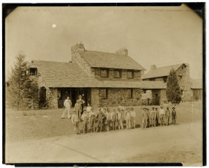 People standing outside of the Chinqua-Penn Plantation Gate House under construction. There is one group on the left, most in working clothes and one standing above the rest in a suit, all of them white. There is another group to the right, all in working clothes and all of them are Black people. The two groups are separated by physical space so they are not standing together, yet still all in the frame.