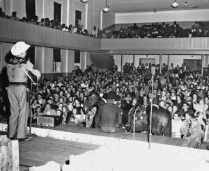 Photo taken from the back of the stage setup in the auditorium. Fred Kirby is performed in a Western stage costume and white ‘cowboy’ hat with embroidery of floral patterns and some beading on the bottom of shirt, belt, and gun holster. Guns with white embellished handles are visible on both sides of his hips. He is playing a guitar made of dark wood and has a strap on. Stage is set up with microphones and 1950’s sound and lighting equipment. An assistant sits on the edge of the front of the stage with Fred’s horse, which has a saddle and reigns on. The floor of the auditorium is filled with white audience members seated for the performance. Above them in a segregated balcony section, all the Black audience members watch the performance.