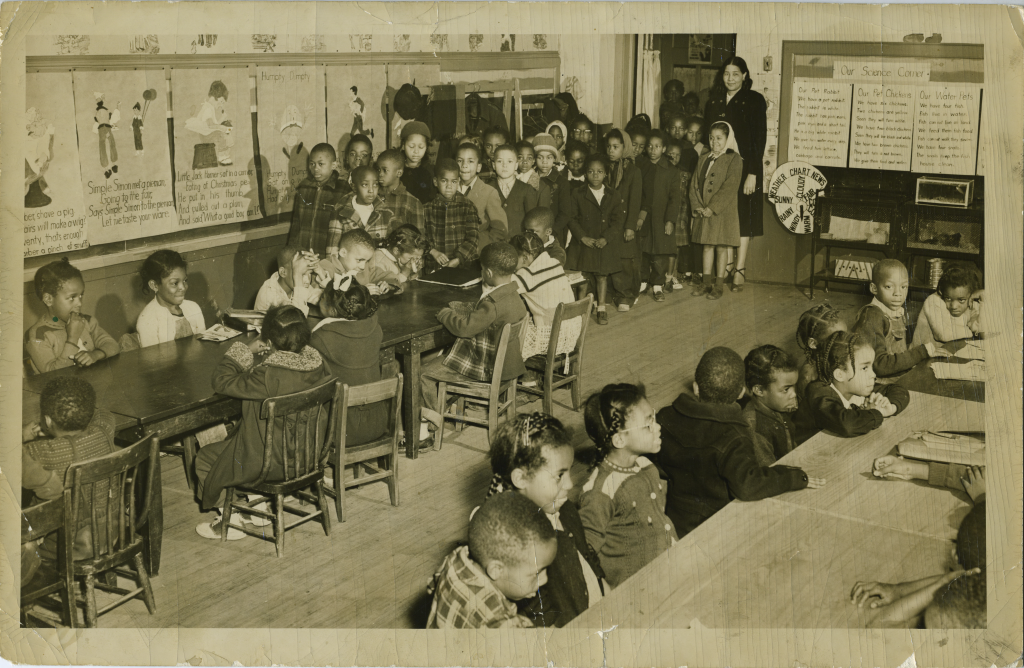 Overcrowded classroom of a Black schoolhouse, two classes in one room, one teacher poses with young children, most of whom are wearing coats, some sitting at the available tables and some standing at the back of the classroom. Classroom is decorated with paper posters and educational materials.