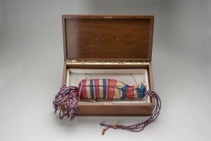 A red and blue bag with tassels that holds a champagne bottle inside. The bag is inside an opened wooden box.