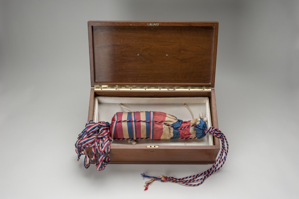 A red and blue bag with tassels that holds a champagne bottle inside. The bag is inside an opened wooden box.