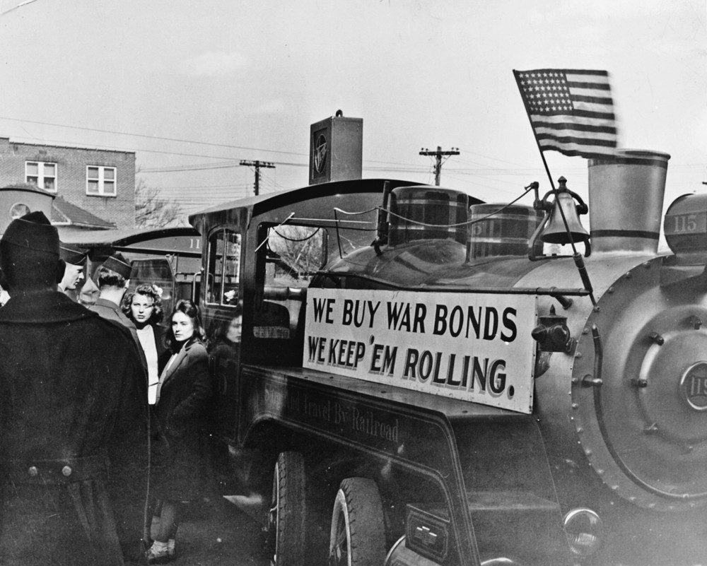 People stand around a train with a sign that reads: "We buy war bonds, we keep 'em rolling." The train flies an American flag.