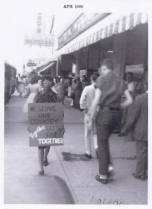 This photo from the collection is taken outside of a F.W. Woolworth location. Features a Black woman with her hair curled and wearing cat-eye glasses and a dress. She is protesting outside of Woolworth holding a sign that reads ‘We serve our country together, let’s eat together.’ People in the street are pictured walking past her. Photo has a date at the top that reads ‘APR 1960’ and is annotated at the bottom with ‘Lofton.’