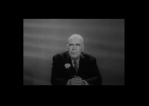 Frame of Former governor Luther Hodges during filmed address to the people of North Carolina about his intention to maintain segregation. The film is in black and white. He appears older and balding. He is wearing a suit and tie with a flower in his lapel. He is sitting during the address, hands folded on top of the desk in front of him.