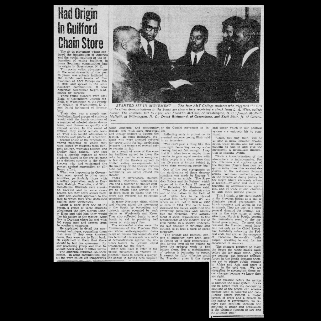 An article clipping from the newspaper on white paper with little damage. The article features a black and white photograph of the four A&T students who began the Greensboro sit-in receiving a check from their college bursar. The students are all wearing suits and ties, one of them with thick black glasses. The bursar is turned towards them, in his suit and tie, presenting them the paper check.
