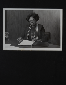 This black and white photo shows Mrs. Millie Veasey sitting at a desk ready to flip through the binder in front of her. There is wood paneling in the background and her desk has papers organized on it. She has her hair curled, is wearing a blouse and jacket with white stripes trimming it, and is wearing jewelry, two long necklaces, hoop earrings, a bracelet and a wedding ring. She is shown wearing aviator-style glasses. The photo is framed with a white border.