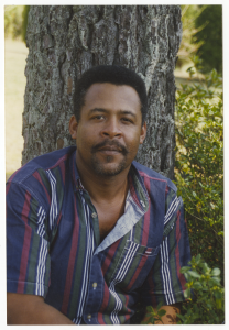 Gregory Tabron is sitting outside in front of a tree. He is wearing a blue, red, green, and white striped button up short-sleeved shirt. He has short, natural hair and a goatee. He has a slight smile for the photo.