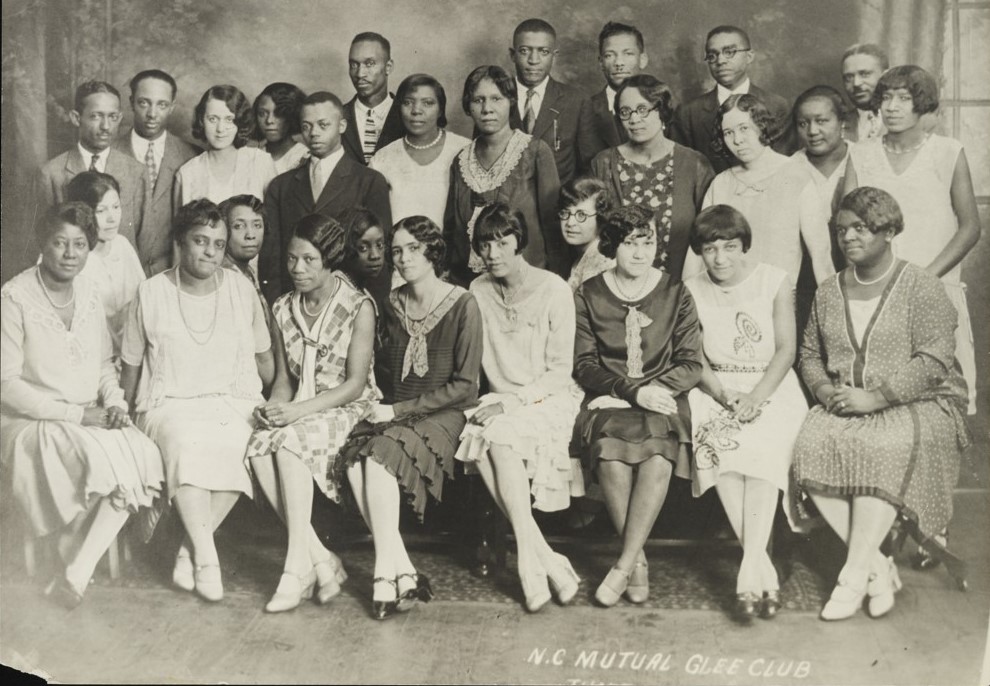 Group of individuals both sitting and standing with the caption "N.C. Mutual Glee Club" written below them. The people are dressed in 1920s attire.