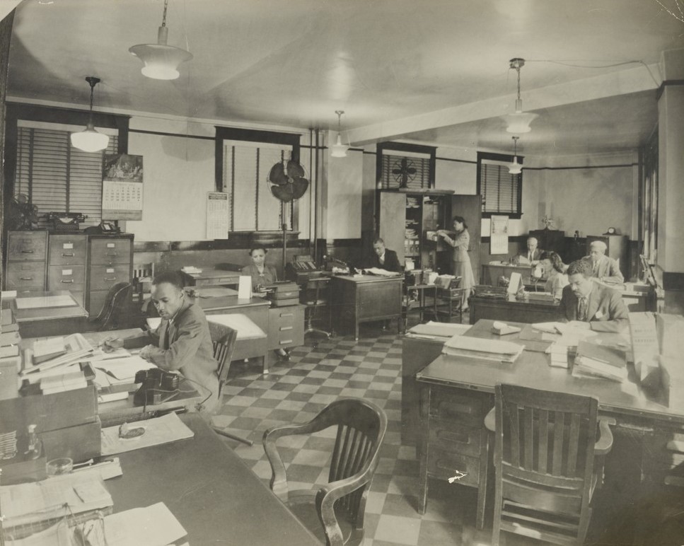 View inside an office. There are numerous desks with papers, books, and other materials on top of them. Several desks have individuals sitting at them working. One person is standing up looking through a filing cabinet drawer.