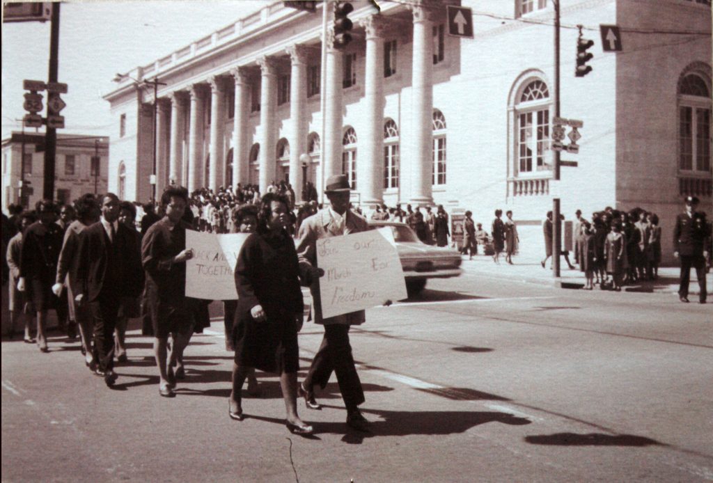 People are shown marching together as a demonstration in the street, in front of a public building. Signs are shown that read ‘Join our March For Freedom’ and ‘Black and W[hite] Together.’ Protesters are wearing business attire and coats.