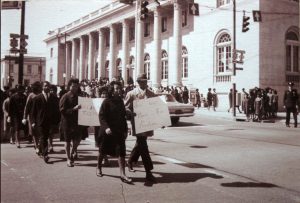 People are shown marching together as a demonstration in the street, in front of a public building. Signs are shown that read ‘Join our March For Freedom’ and ‘Black and W[hite] Together.’ Protesters are wearing business attire and coats.
