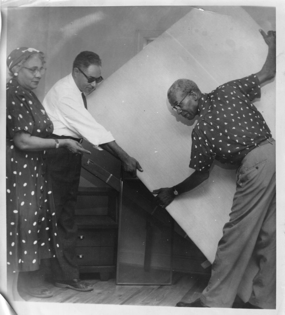 A snapshot showing a man holding a large object of furniture and moving it with the assistance of another man. To the left of them, a woman is pointing and appears to be directing the moving effort. 