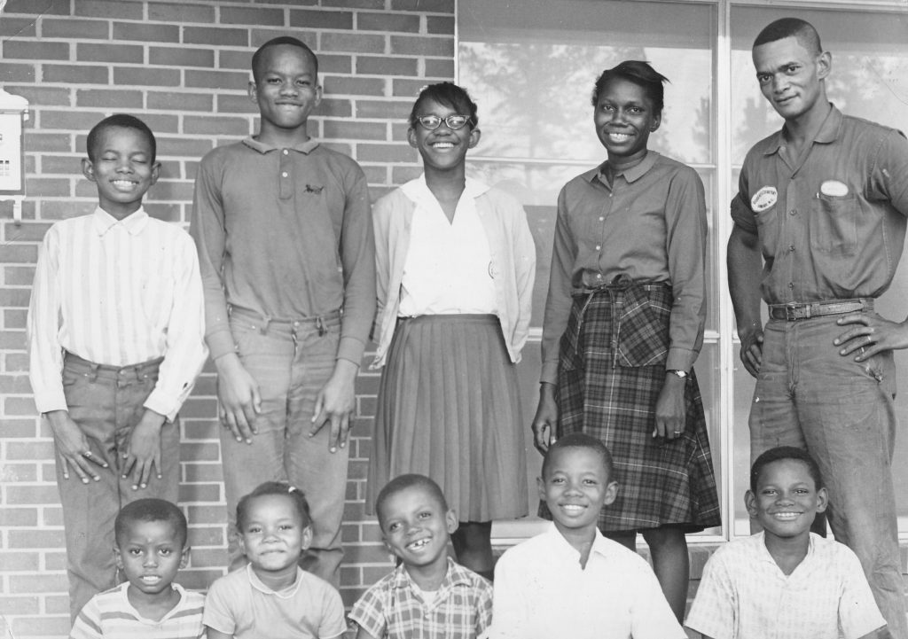 A photograph of a smiling family on the porch of their new home. The parents of the Womack family stand to the right of the frame, alongside three of their children. Directly below and in front of them is a row featuring five sitting children.