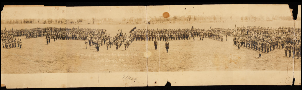 Part of a panoramic photograph that depicts military troops organized in a special formation across a large field. The troops are dressed in official military uniforms and many individuals hold flags. Numerous lines of handwriting say, "Memorial Ceremonies for Pres. Harding", "Fort Bragg N.C.", and "August 6, 1923"