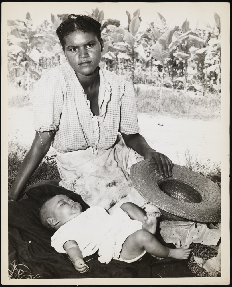 Woman and baby on a blanket next to a field of tobacco. The woman is sitting upright on the blanket with a hat in her hand. The baby is laying down on the blanket.