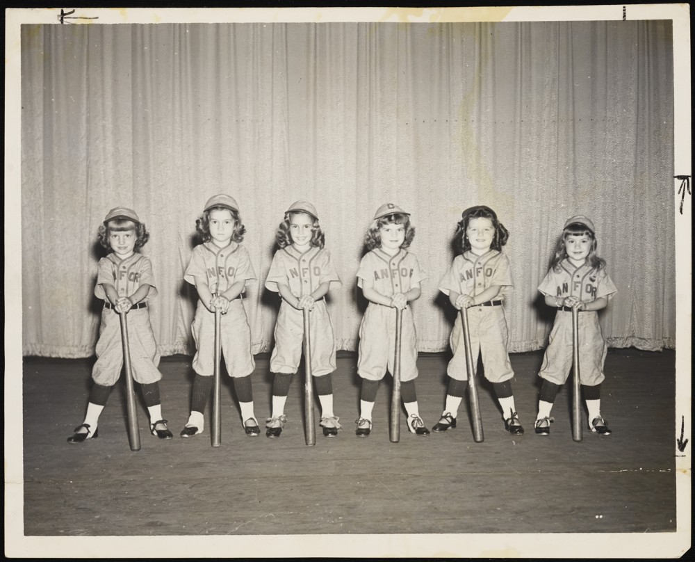 Six children lined up side by side wearing softball uniforms including a hat, jersey with "SANFORD" written across the front, pants, and cleats. In front of them they have softball bats.