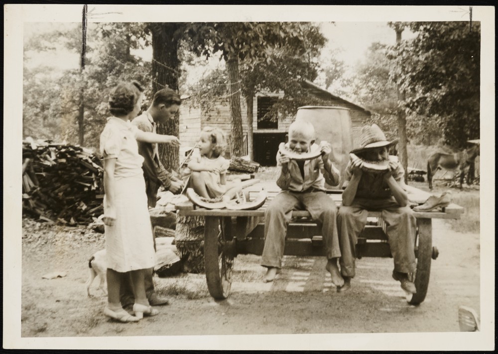 Two adults and three children. The three children are sitting on the back of a trailer. The two children to the right have their legs hanging off the back of the trailer and are eating wedges of watermelon. The third child, sitting a little further back on the left of the trailer, is looking at one of the adults with two eaten watermelon edges in front of them. The two adults are on the left side of the trailer. The one in front is wearing a light colored top and skirt and has their arm held out towards the back of the trailer. The second adult stands behind the first adult potentially chopping wood.