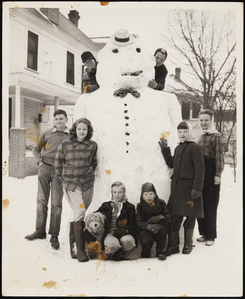 Eight children standing around a giant snow man. In relation to the snowman there are two children to the left, two children to the right, two kneeled in front, and two standing behind (presumably standing on top of something).