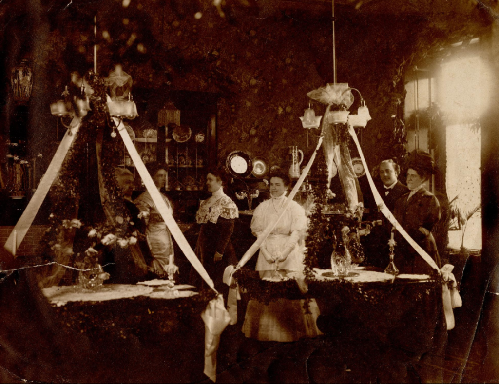 This sepia-toned photograph shows several people dressed in formal attire standing behind tables that are intricately decorated for an event. 