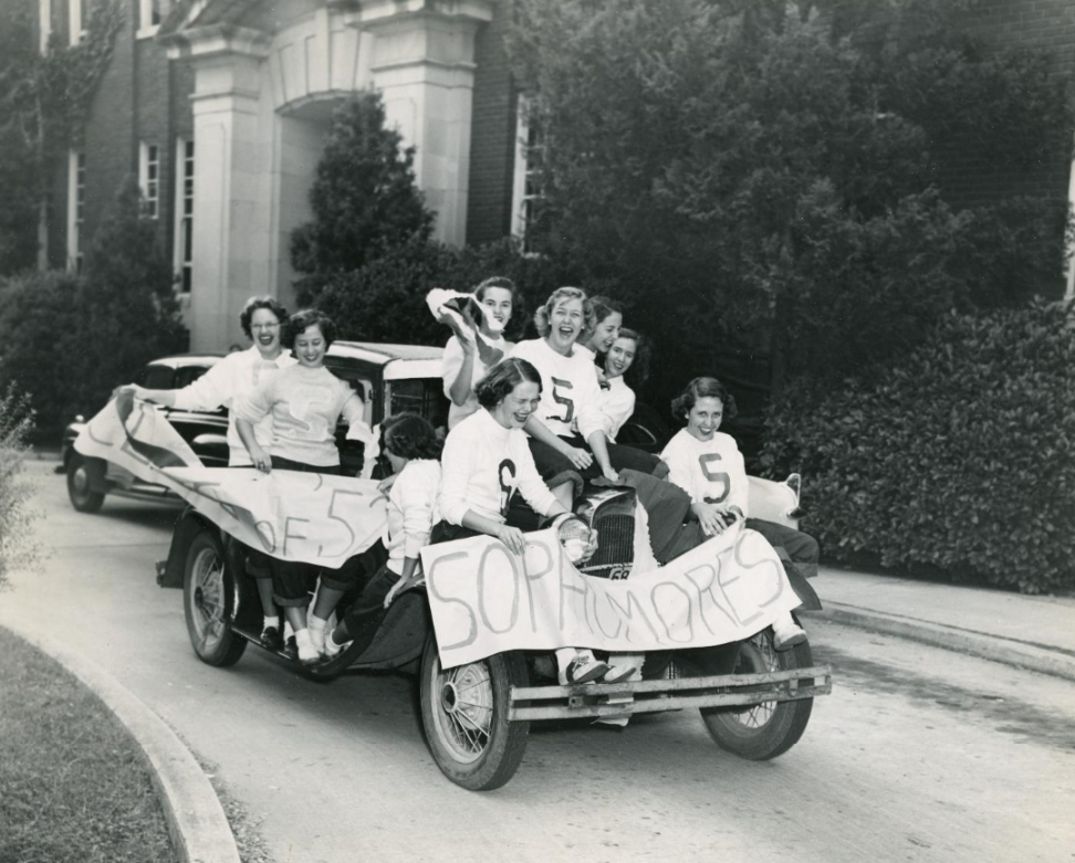 Nine college students sit inside and on top of a convertible car while holding two white banners that read "SOPHOMORES" and "CLASS OF '52"
