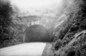 The entrance of a stone tunnel, surrounded by trees, shrubbery, and a rocky mountainside. A paved road runs through the tunnel.