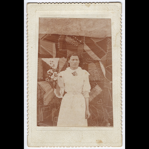 This sepia colored, possibly tin-type, photo has a cream scalloped border. Ormand Goforth is standing in front of her quilt, hair pulled back and neutral expression. She is looking off to the side and wearing a white dress that has a white belt cinching her waist and elbow length sleeves which have a ruffle of lace extending past her elbow. There is a flower brooch pinned at her collar, which is lace that fall like a turtleneck neckline, and lace adorns the shoulder and chest area of the dress. She is holding a small bouquet of flowers up by her shoulder for the camera. Her quilt is hung behind her so the full length can be seen by viewers at the event. In the photo, you can see the asymmetrical pieces, all various sizes, though not many are extremely small. Different shades can be seen and some fabrics used have patterns themselves and others are solid color. On top of the stitching for the quilt, she has added more visible stitching, likely embroidery style extenuating the asymmetry and shapes of the pieces of fabric, some in zigzags, others in x patterns, others in a running stitch. She has also stitched in her initials, what seems to be the beginning of her date of completion, a branch of leaves, and a heart with double lines visible in the photo.