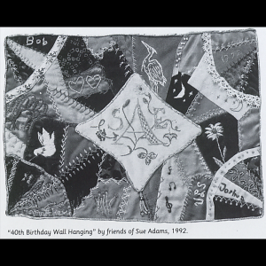 Quilt made in the crazy quilt style; it has many small pieces of fabric (most solid colors) sticked together with a large diamond shape white piece of fabric in the middle. Inside the diamond is this stitched initials of the recipient ‘SAE’ and small floral accents added around the initials; throughout the quilt there are the names of contributors stitched into the quilt as well. There are many visible embellished stitches on top of the regular stitches, as well as lace and embroidered/stitched designs like flowers, moon, stars, dove, crane, hearts, music notes, and a butterfly.