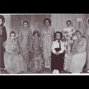 9 women are posed for a picture, staggered between standing and sitting. The four women in front sitting all have their hands crossed in their laps. The five women standing in between those sitting have their hands by their sides and the two women on the ends of the photo are partially out of the frame. They are all dressed in traditional circa 18th century clothing, akin to typical Amish attire, though most of their dresses are made with floral or patterned fabric and lace accents. All are wearing bonnets and smiling for the photo. The background is a wood paneled wall, and electrical wiring can be seen running into the wall.