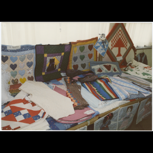 Pictured is a table filled with the class’s quilting projects. Hanging in a row above the back of the table are five completed quilted squares, two have applique multicolored hearts, one with three rows of three hearts and the other with four rows of four hearts. Another features a black cat applique with a little purple bow stitched onto the cat. Another has a geometric pattern of white and blue triangles arranged to look like squares. The last is a geometric design with a large triangle that has an attached rectangle and trapezoid to mimic a trunk and base of a tree, then has small triangles arranged around the larger triangle to mimic leaves. On the table are piles of quilted projects, some barely visible in the photo. There are pieces with applique flowers, geometric patterns, a single piece of patterned fabric for the top layer, and thin stripes of different colored material stitched together for the top layer of the quilted project. There is a piece that also appears to be some kind of garment meant to be worn, perhaps the beginning of a shirt or apron. Hanging along the front of the table are squares with a person (what might be representative of a young girl) appliqued onto the square, one seeming to have used fabrics to represent darker skin and curly hair with a bow and a blue and red dress, another uses fabric to represent very light skin, brown short hair, and a pink dress.