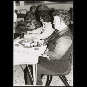 In the forefront, a woman wearing glasses and dressed in a collared shirt, sweater vest, and pants looks up while she is working on a quilt square held in a quilting frame. She is sitting at a table and stitching an applique design onto the fabric in the square. In the background, other students are sitting around the table and many are writing, presumably planning designs or completing work for the class.