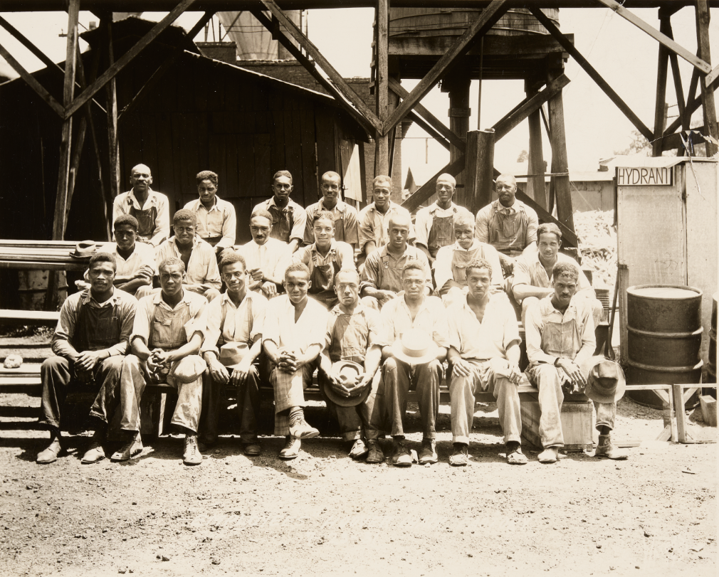 22 men sit in three rows on bleachers in the outdoor area of an industrial factory.