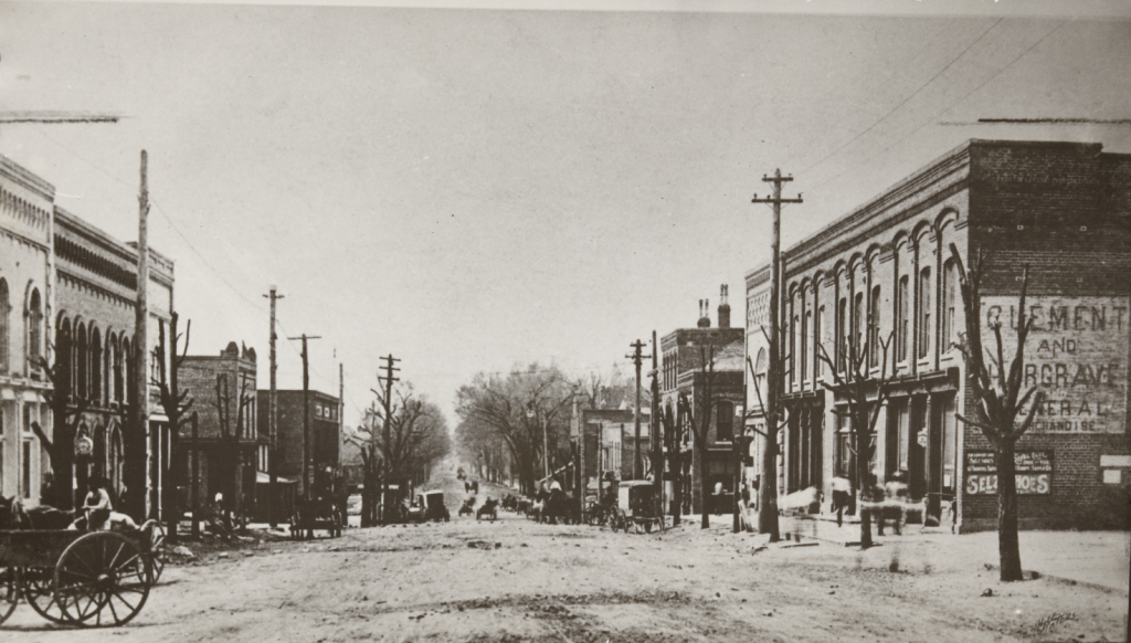 A view of South Main Street in Lexington, N.C. from around 1910. The black-and-white photograph shows dirt roads lined with brick stores and telephone poles. Several horse-drawn wagons can be seen parked and moving along the street.