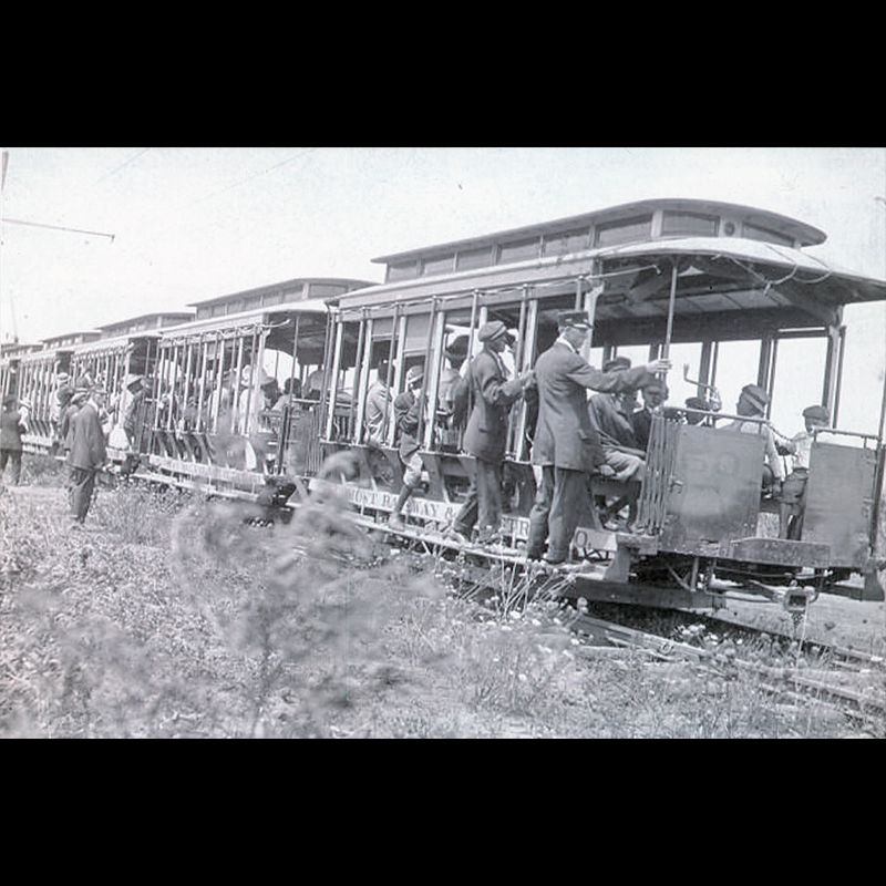 Photo of streetcars with multiple cars connected together. People are filling the cars and there are two individuals wearing sites and hats visible standing off the the side observing the streetcars and passengers while two other individuals also wearing suits and hats are hanging off the side of the first streetcar in the photo.