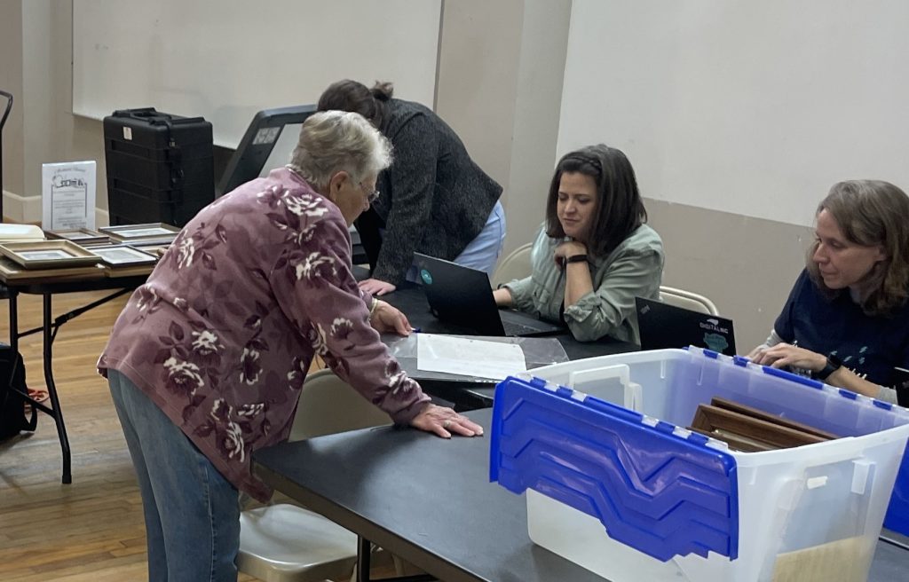 Two individuals talking. The person on the left is pointing to an object on a table while the person on the right is working on a laptop.