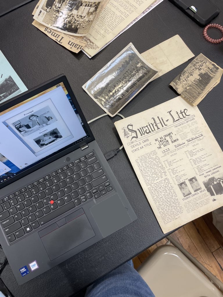 A table that has a laptop, newspaper, and photographs on it.