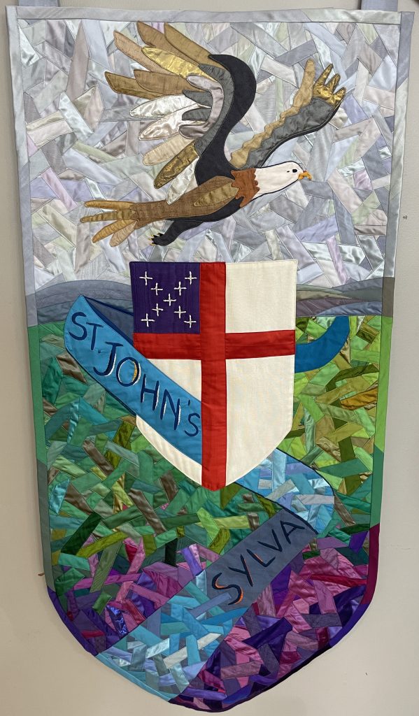 A quilted banner with images of a shield or coat of arms, bald eagle, and a ribbon with the text St. John's. Sylva.