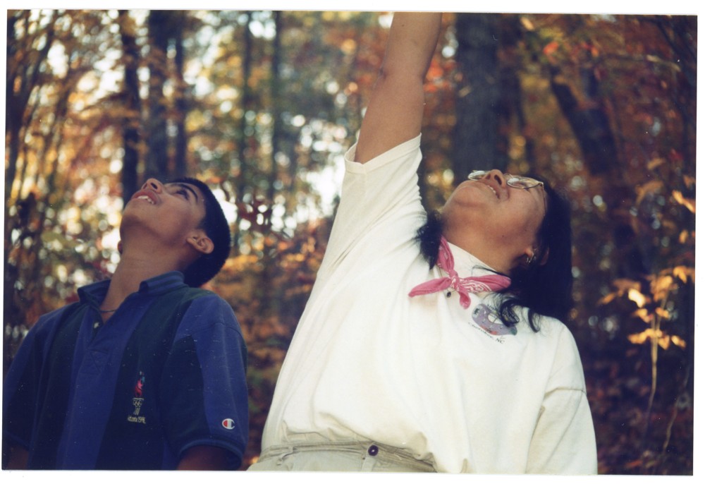Two individuals in a forest looking upwards. The person on the right has their hand raised most likely to point something out.
