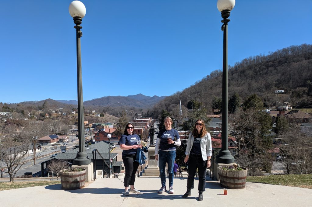 Three individuals wearing the same t-shirt standing on top of a hill. Behind them is a town and mountains.