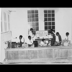 Eight people are standing behind a long work counter, some working on various radio parts distributed across the work counter and others studying an informational poster titled “Dynamic Demonstration.”