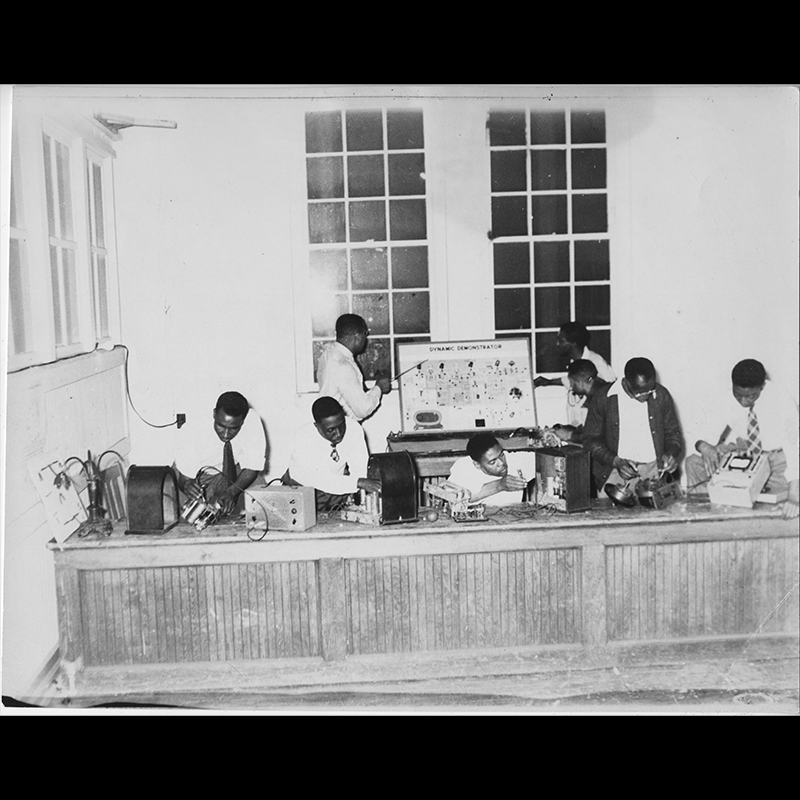 Eight people are standing behind a long work counter, some working on various radio parts distributed across the work counter and others studying an informational poster titled “Dynamic Demonstration.”