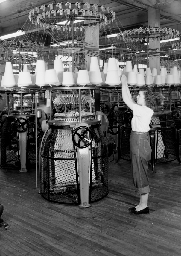 A woman at Barbet Mills reaches up to adjust textile spools that are suspended from a large machine.