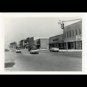 Black and white photography of Downtown Kings Mountain, seen down Battleground Ave. The McGinnis department store can be seen, as can several other business signs, the last building seen is the First Union Bank building. Five white light posts that can be seen running along the road in front of the buildings. Multiple cars can be seen on the road, some are driving up the road, others are driving down the road toward the photographer. A stop light can be seen on the top right side of the image.