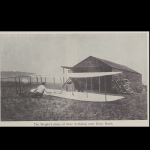 Photo of Orville and Wilbur Wright working on their first plane, one squatting and reaching across the cockpit space and the other leaning against the top of the wing on the opposite side. The workshop barn is seen in the background.
