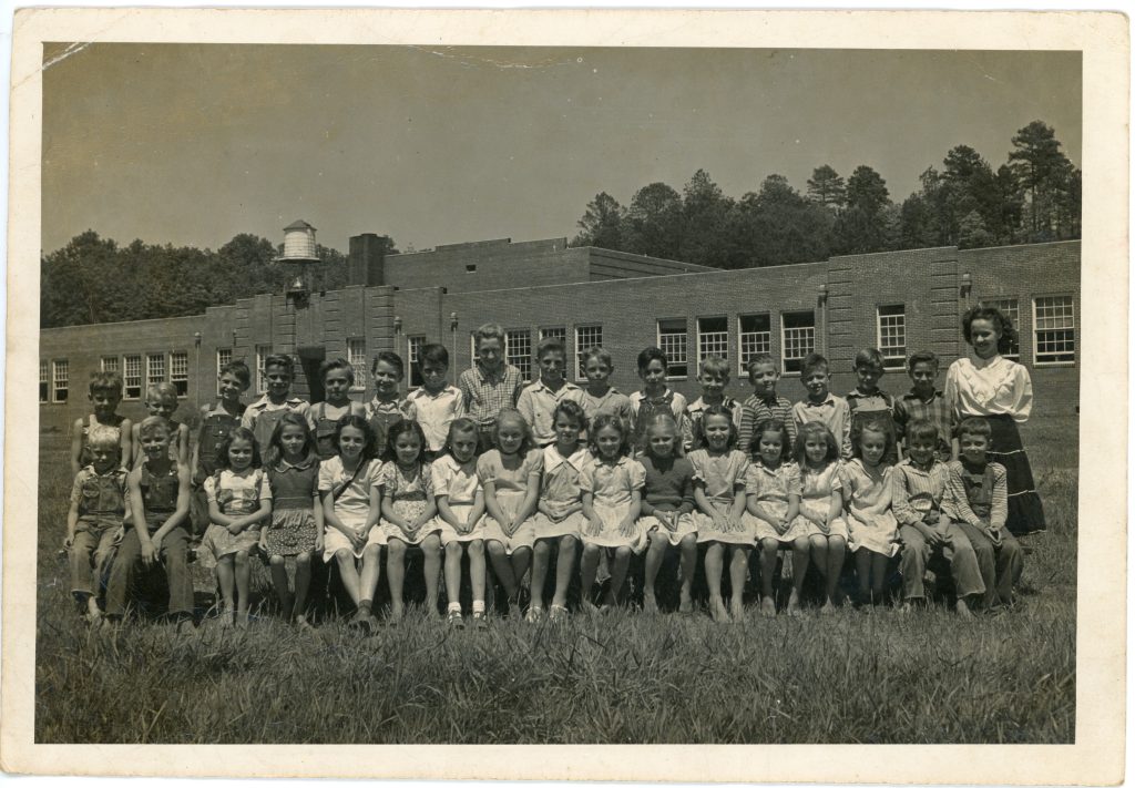 Two rows of first grade aged children. The front row is seated on a bench while the back row stands. On the far left is the teacher.