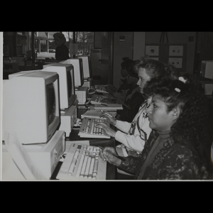 Computers are lined up on tables and 4 are visible in the photo with a student at each computer. They are all focused on their computers and some are taking handwritten notes.