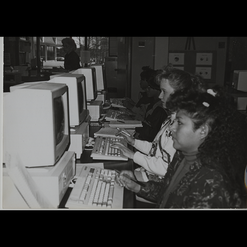 Computers are lined up on tables and 4 are visible in the photo with a student at each computer. They are all focused on their computers and some are taking handwritten notes.
